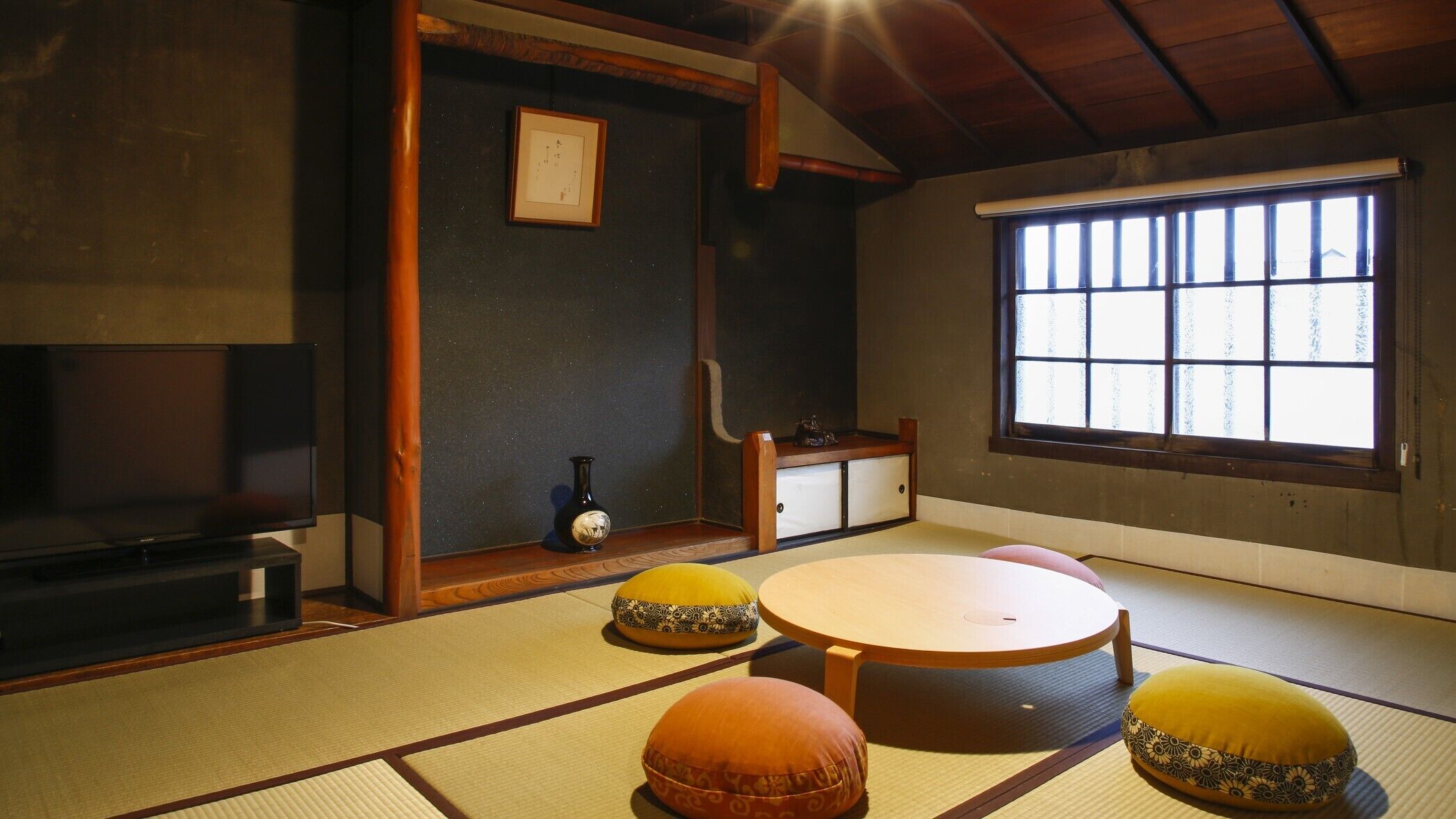 Ein Zimmer mit Tatami-Matten, einem Tatami-Tisch, Kissen, einem kleinen Schrank und einem Fenster mit Aussicht.