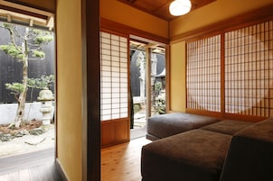 Una habitación japonesa tradicional con suelo de tatami, una cama de madera baja, puertas correderas y vista a un jardín con un árbol y un farol de piedra.