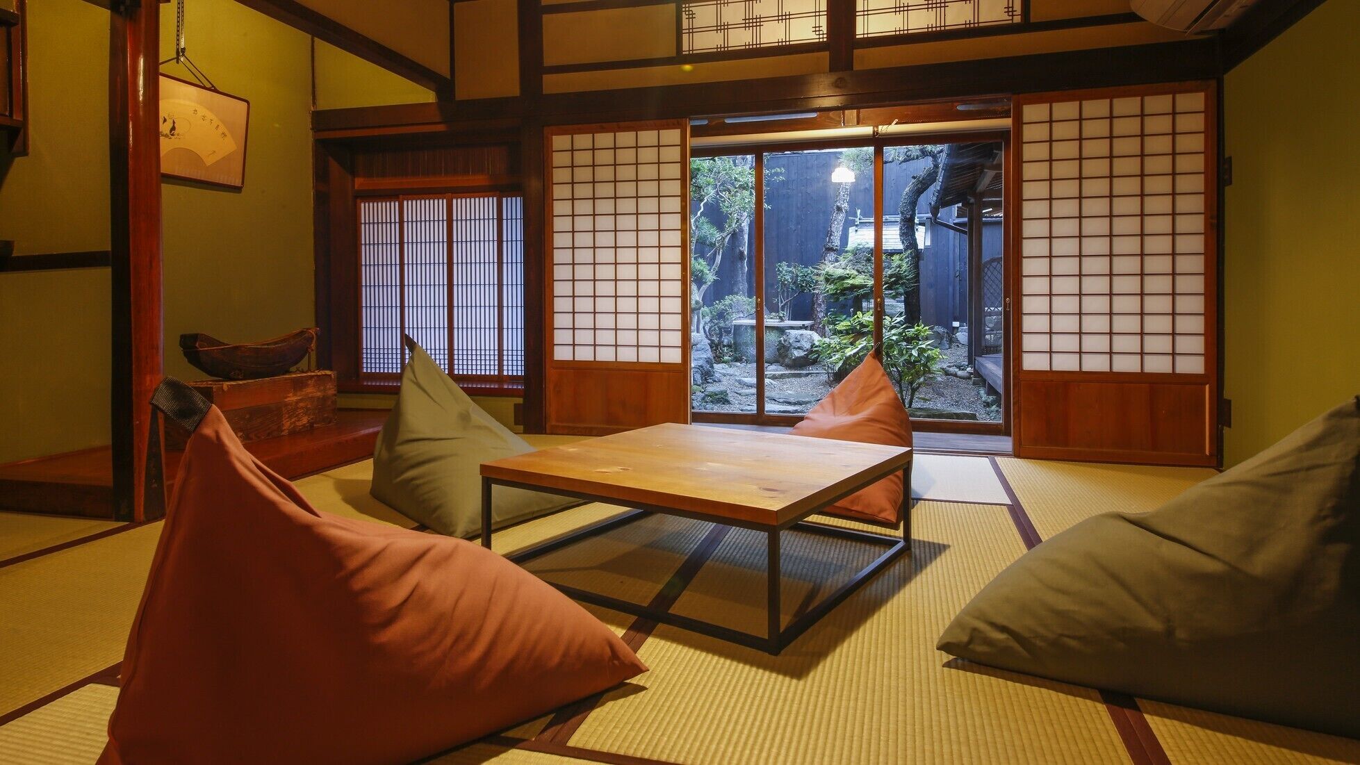 Ein traditioneller japanischer Raum mit Tatami-Boden, einem niedrigen Tisch, Kissen und verschiebbaren Türen, die zu einem Garten führen.