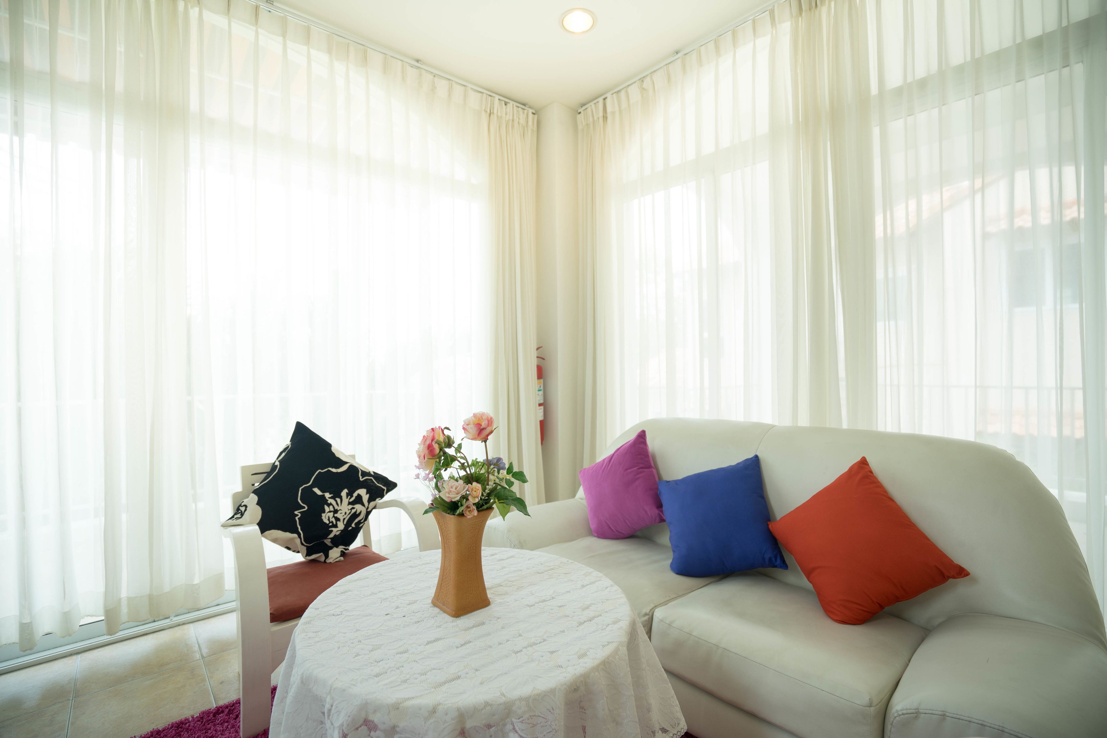 A living room with a white sofa, a round table with a vase of flowers, and sheer curtains.