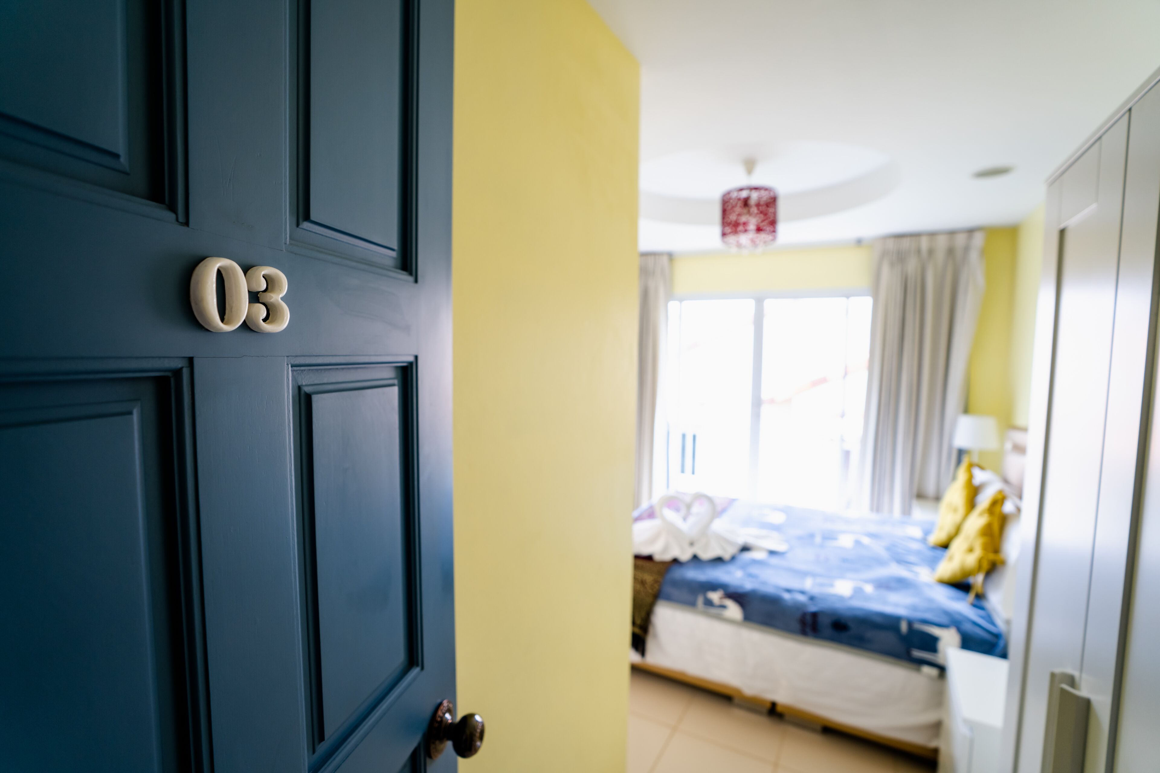 A room with a closed door marked with the number 03, a bed with a blue bedspread, and a window with curtains.