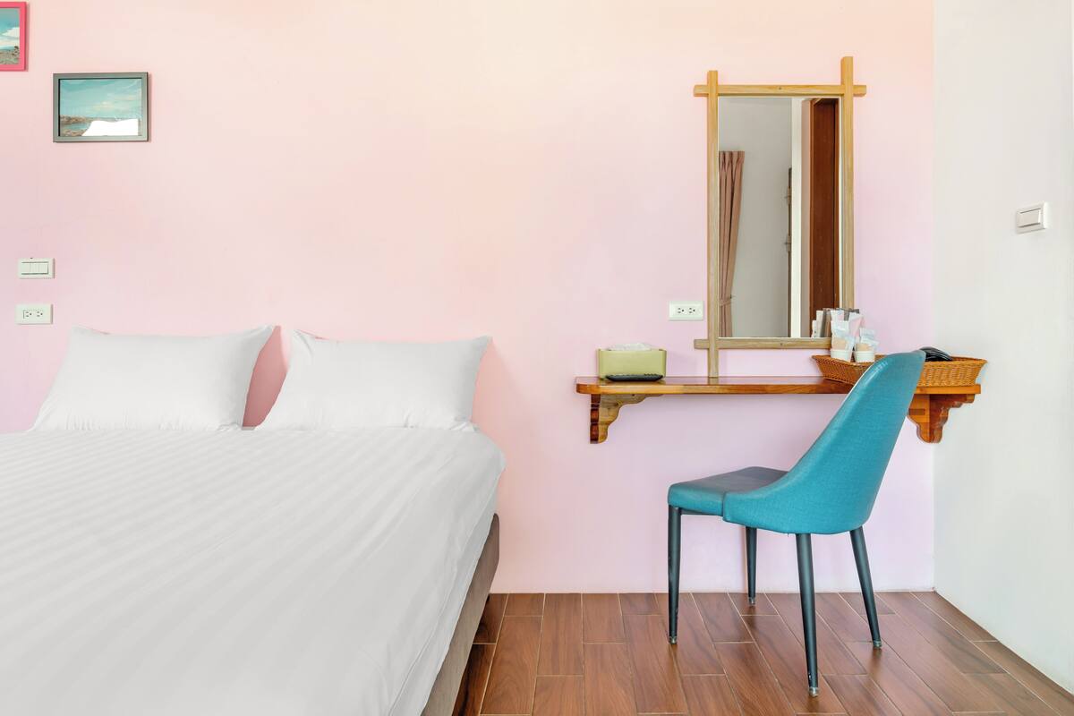 Un dormitorio con cama, una silla, un estante de madera con un espejo y una mesita.
