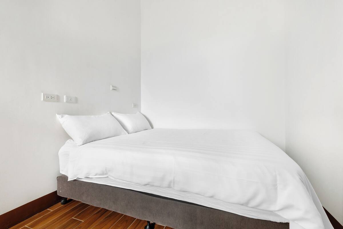 Una cama individual con ropa blanca en una habitación de paredes blancas y suelo de madera.