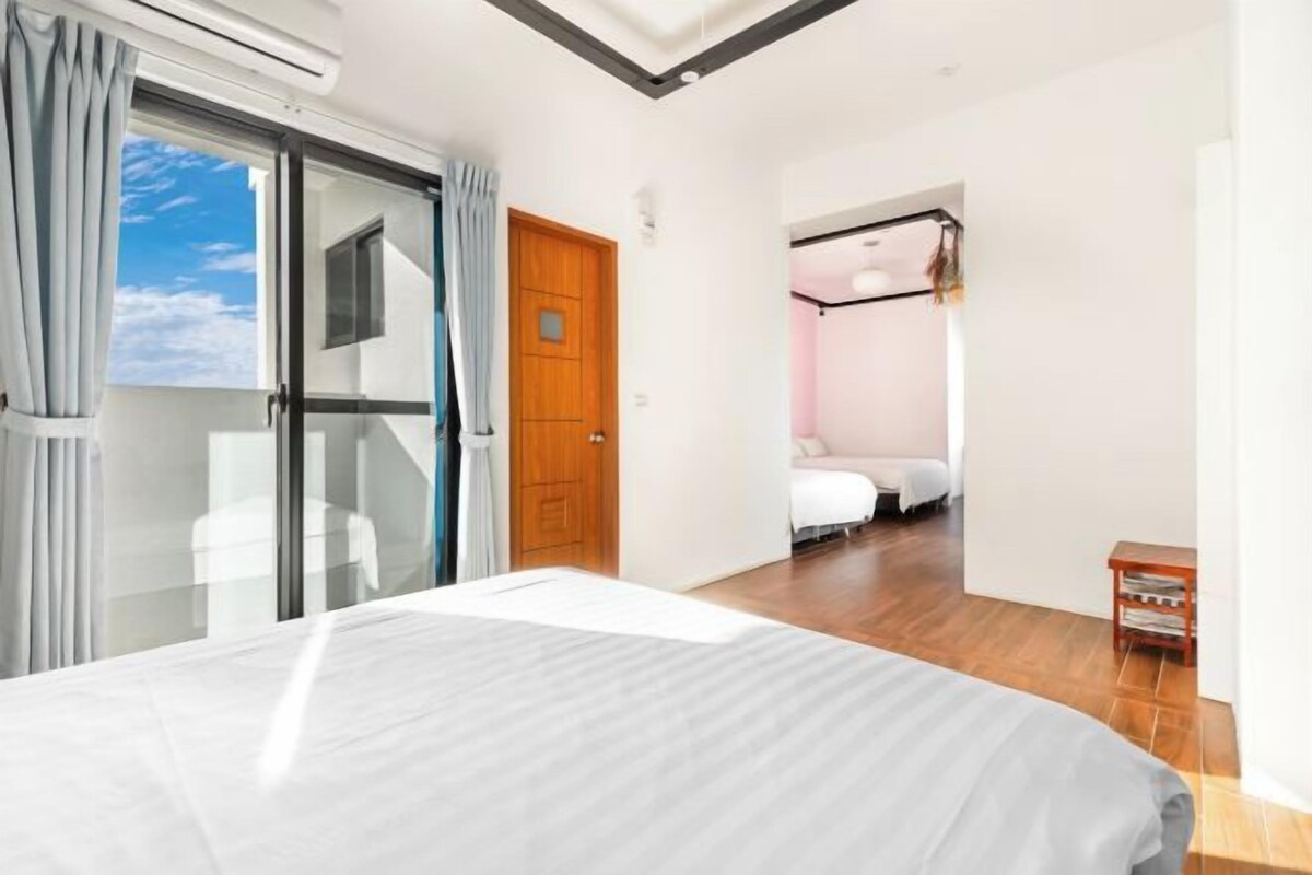 A modern hotel room with a large bed, a sliding glass door leading to a balcony, and a separate bedroom with a bed and a desk.