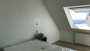 Huone, jossa on kalteva katto, kattovalaisin, sängyssä patjansuojus, patteri ja pieni pöytä, jonka päällä on muovipussi.