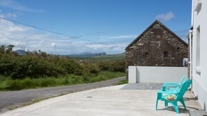 Un edificio de piedra con techo inclinado, un área pavimentada con dos sillas turquesas y vistas a colinas distantes bajo un cielo despejado.