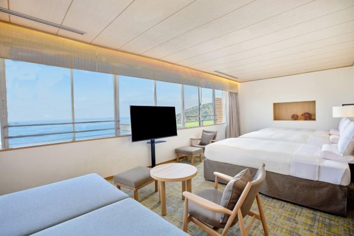 A hotel room with a bed, a TV, a sofa, a chair, a small table, and a view of the sea.
