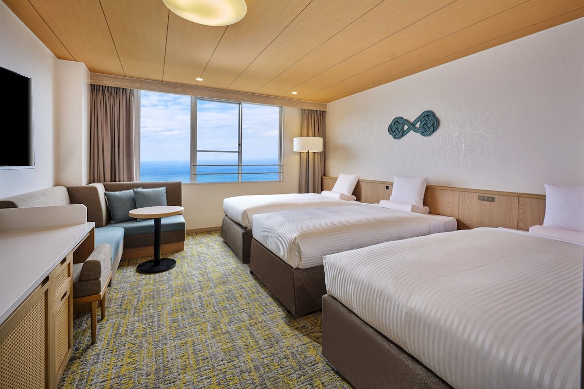 A hotel room with two beds, a sofa, a small table, and a view of the sea.