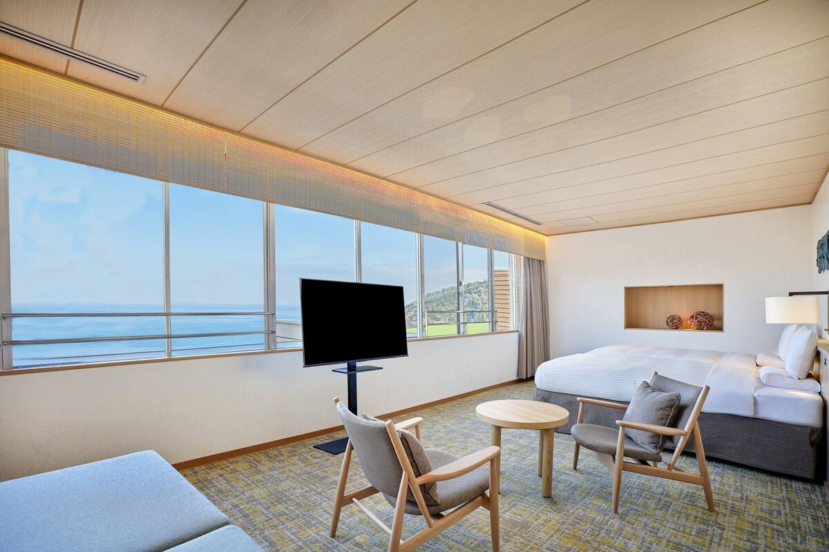 A hotel room with a bed, a TV, a sofa, a chair, a small table, and a view of the sea.