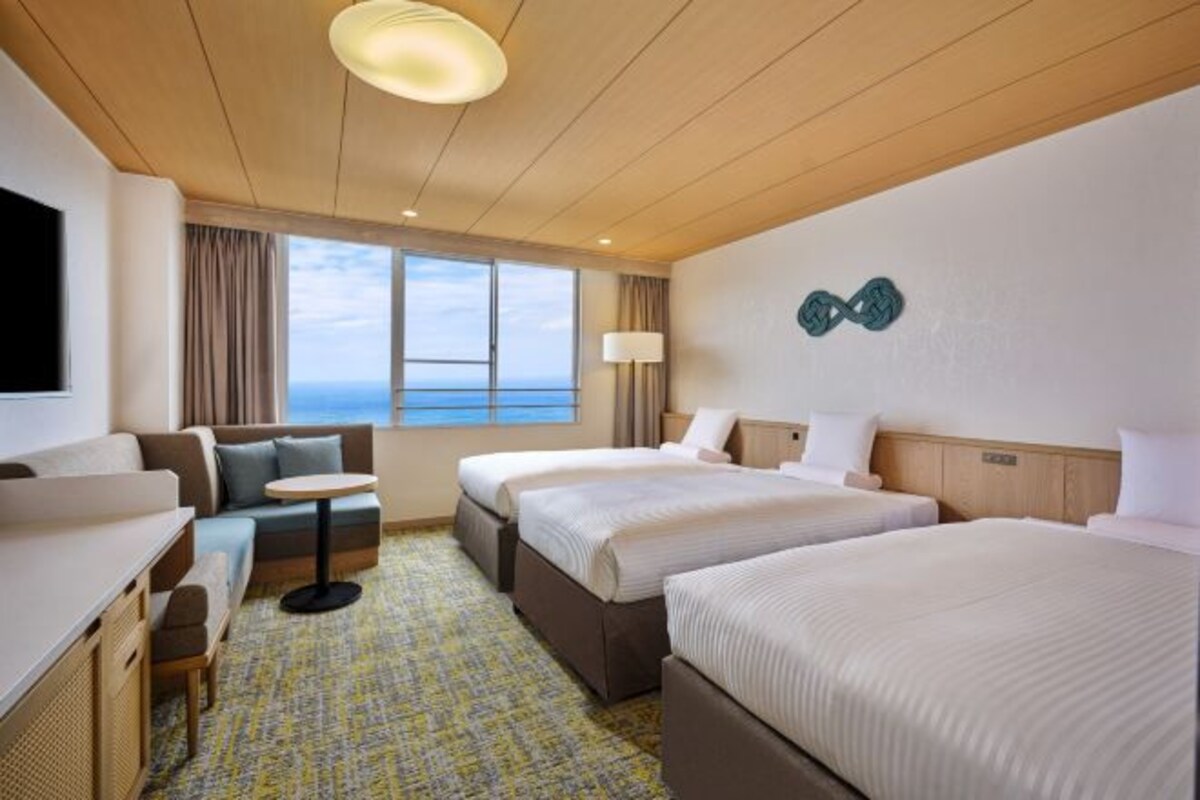 A hotel room with two beds, a sofa, a desk, a small table, and a view of the sea.