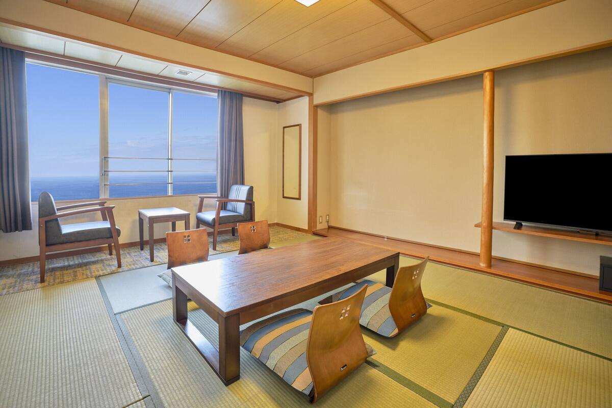 A hotel room with a large wooden table, chairs, a television, and a view of the ocean through large windows.