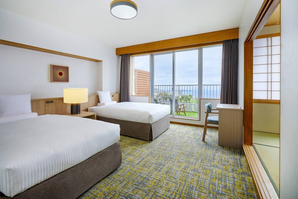 A hotel room with two beds, a desk, a chair, a large window with a view, and a ceiling light.