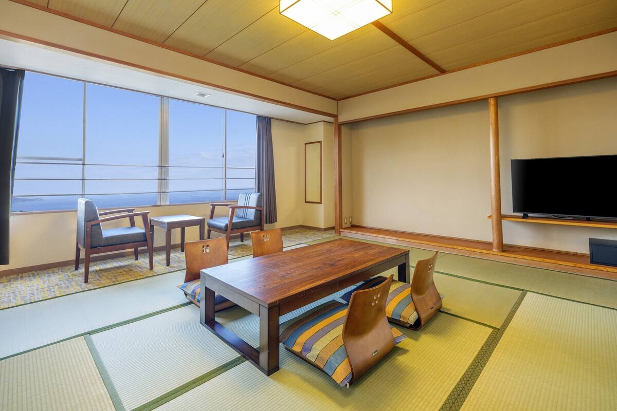 A room with tatami mats, a wooden table, chairs, a television, and a large window with a view.