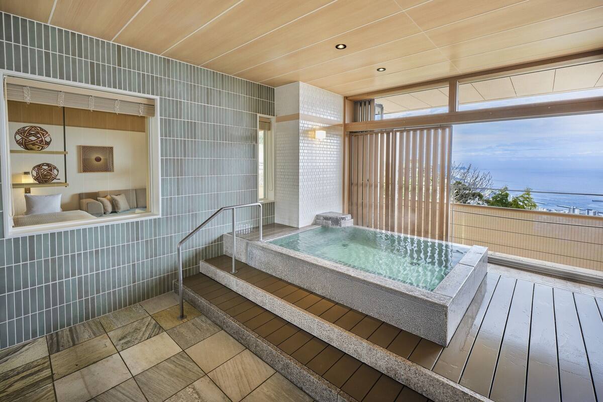 A modern bathroom with a glass-enclosed jacuzzi, a seating area with cushions, and a view of the sea.