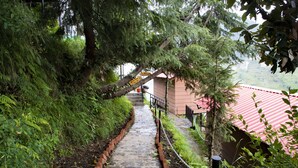 Un chemin de pierres mène à un bâtiment surmonté d’un toit rouge, qui est entouré d’une végétation luxuriante et se trouve au pied d’une pente abrupte.