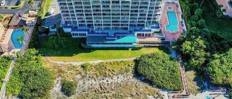 A multi-story hotel with a pool area, surrounded by greenery and a beach.