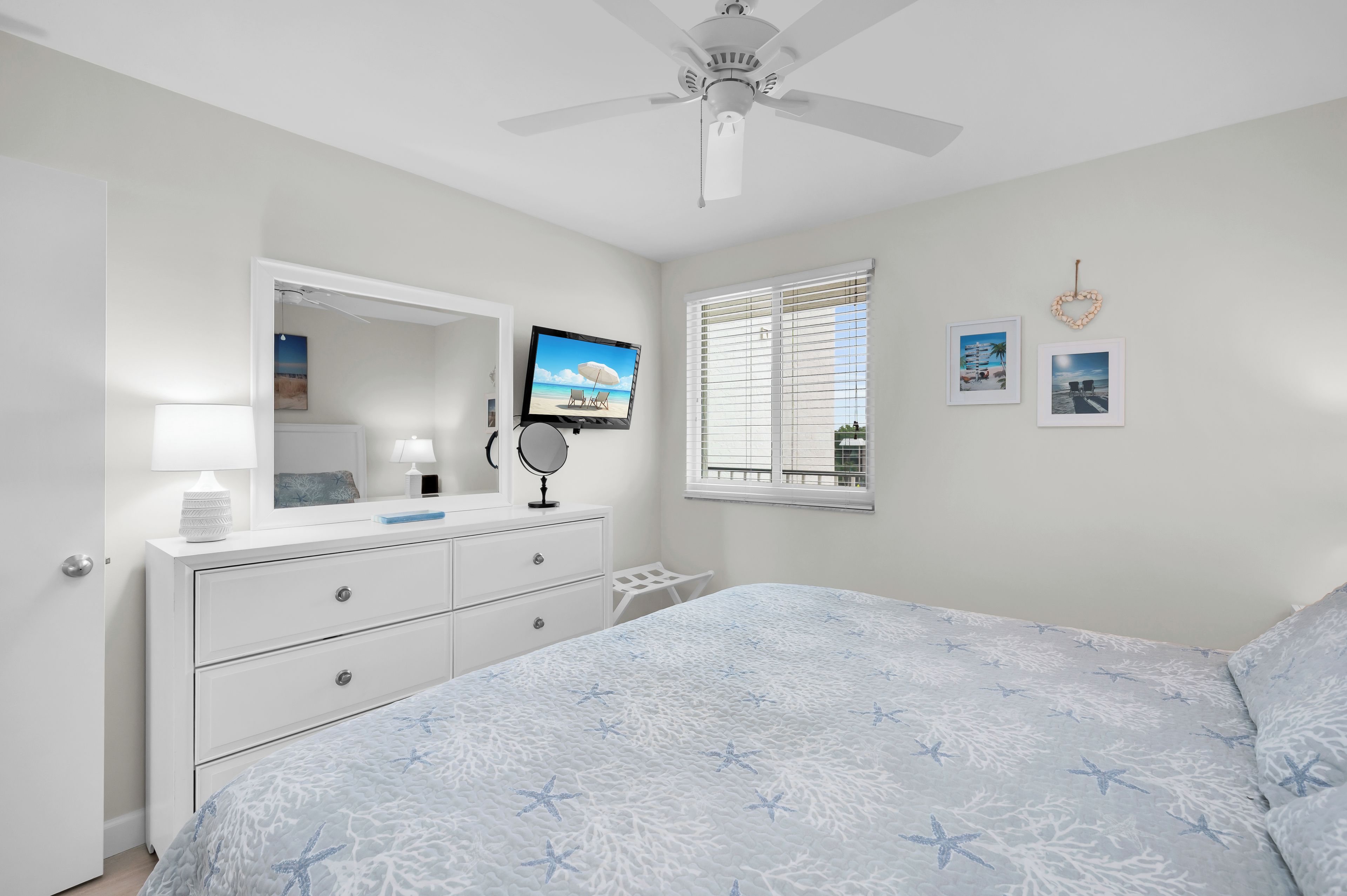 Un dormitorio con cama, tocador, televisor, ventilador de techo y ventana con persianas.