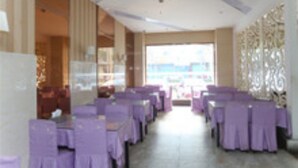 A restaurant with rows of tables covered in purple tablecloths, a large window with a view, and a decorative wall on the right. - Gang Wan Yin Xiang Hotel (Jiaxing)