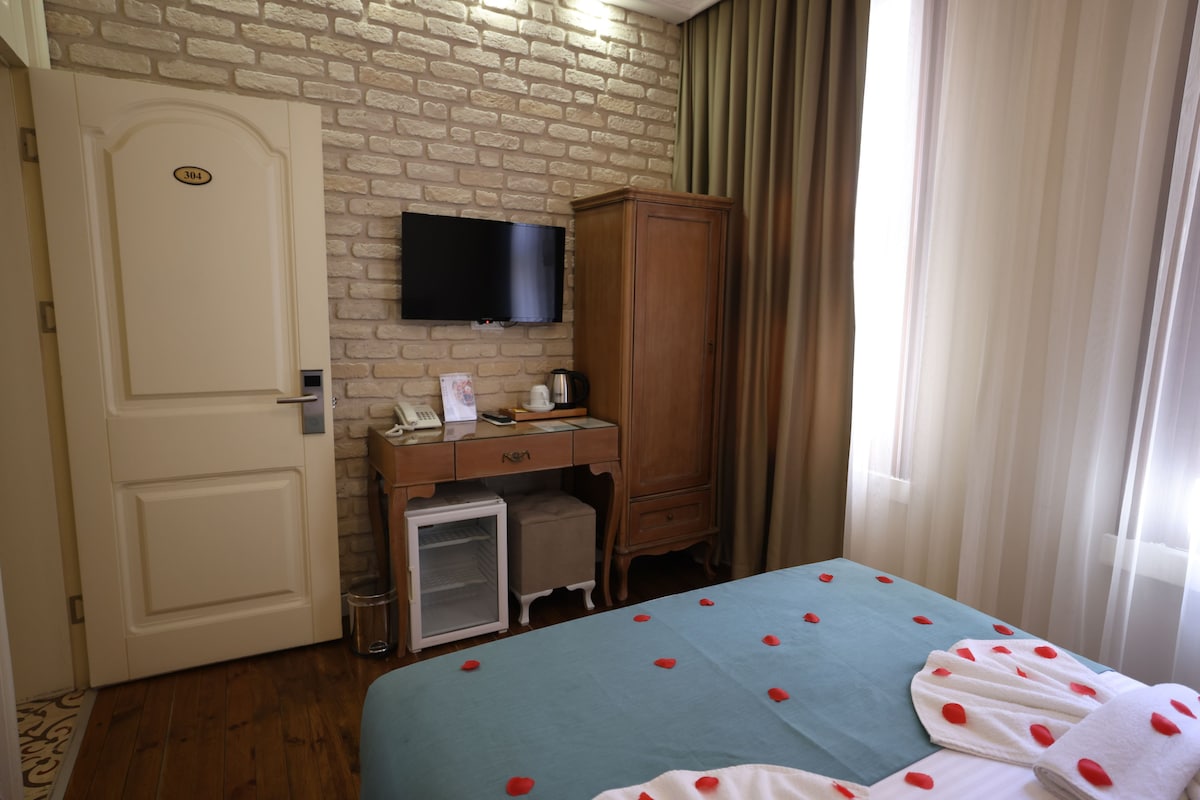 A hotel room with a brick wall, a television mounted above a wooden desk, a bed with rose petals, and a door with a number.