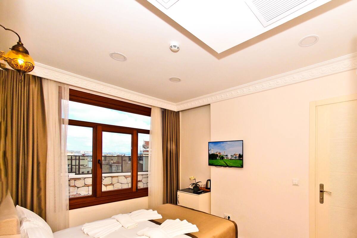A hotel room with a bed, a window with curtains, a red rug, and a painting on the wall.