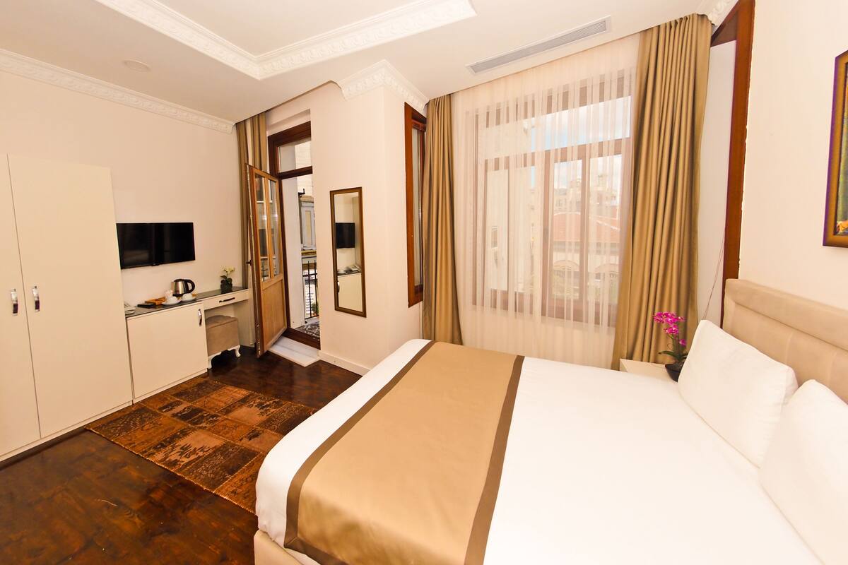 A hotel room with a large bed, a desk with a flat-screen TV, a mirror, and a view of the city through sheer curtains.