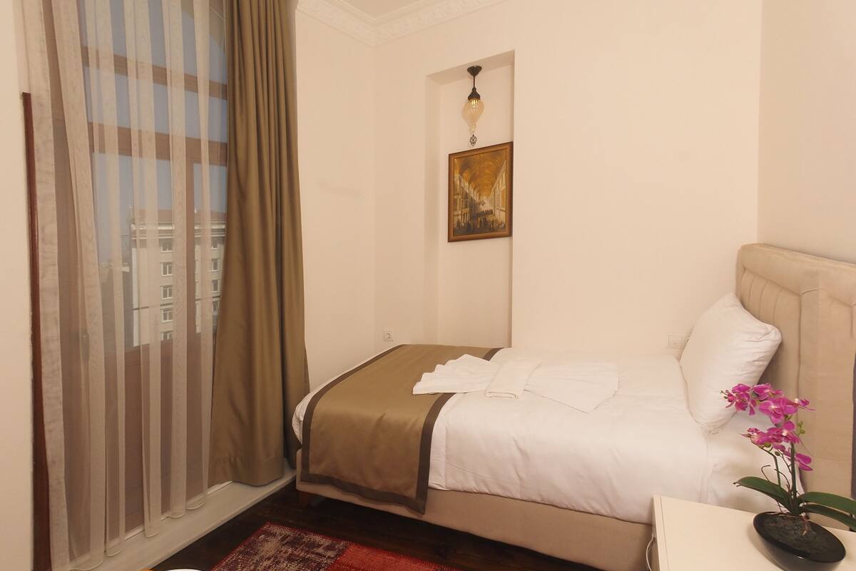 A hotel room with a bed, a desk with a plant, a fruit bowl, and a window with curtains.