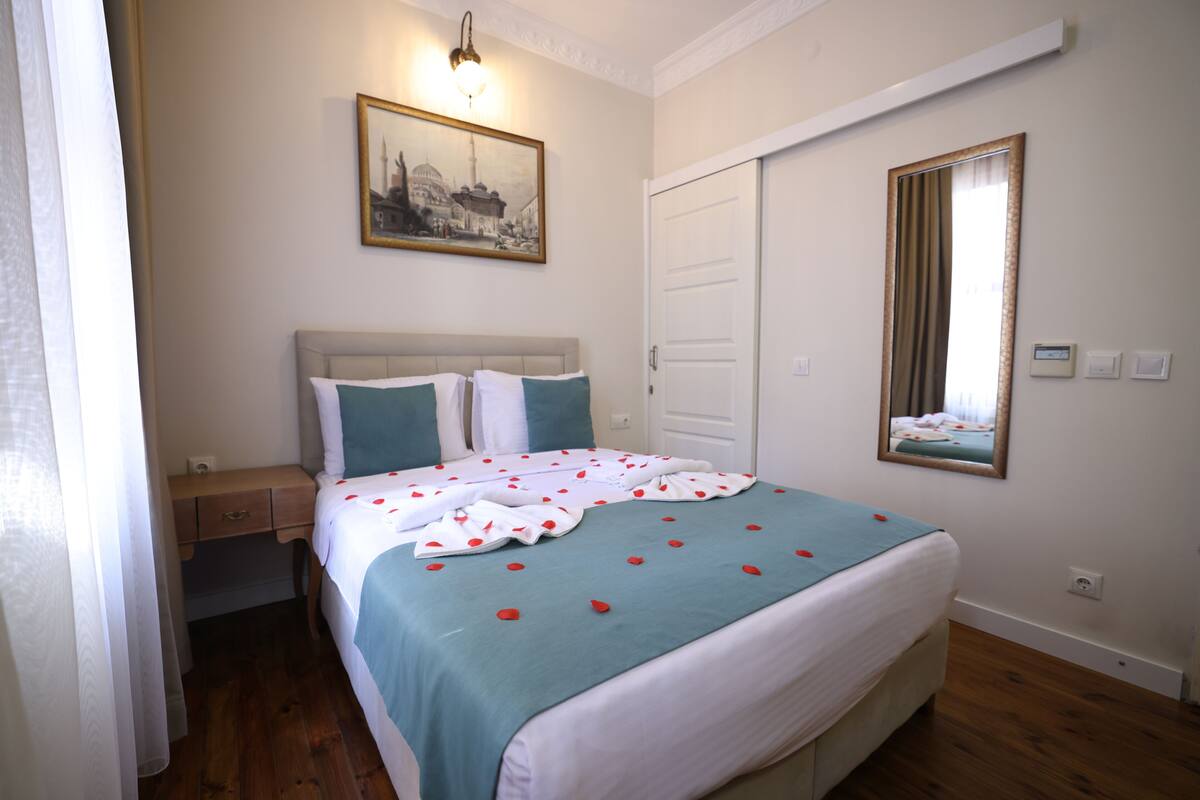 A bedroom with a bed, a nightstand, a bedside table, a wall-mounted painting, a door, and a mirror.