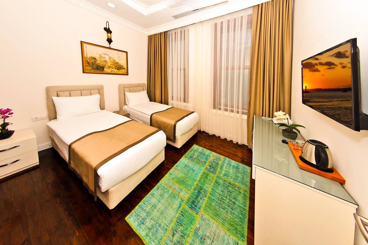 A hotel room with two beds, a large window with curtains, a flat-screen TV, a small table with a kettle, and a colorful rug.