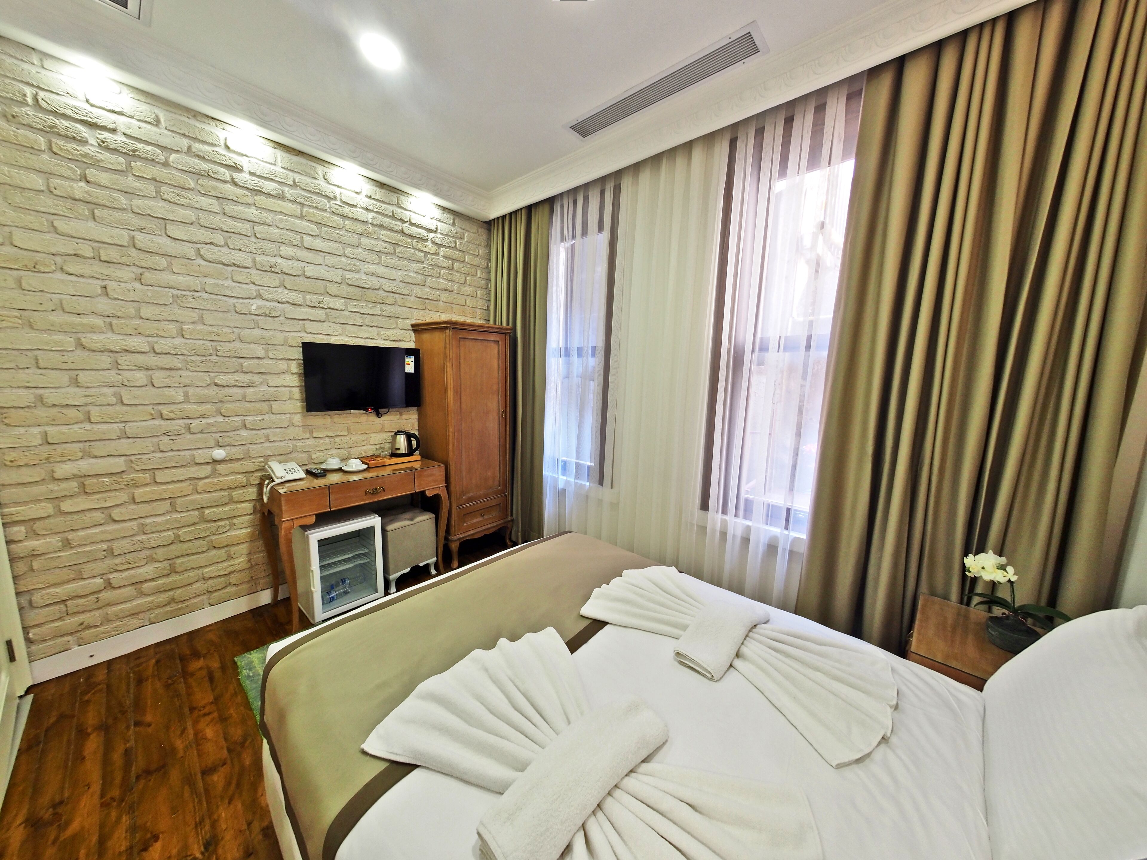 Een hotelkamer met een wand in baksteenmotief, een televisie, een houten kastje, een kleine koelkast, een bed met witte lakens en een raam met gordijnen.