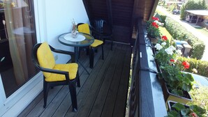 Une terrasse en bois avec deux chaises jaunes et une petite table, donnant sur un jardin agrémenté de plantes en pot et offrant une vue sur la rue.