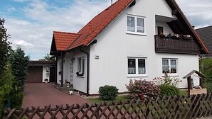 Une maison à deux étages, avec un toit recouvert de tuiles rouges, un balcon à l’étage supérieur et une clôture en bois dans le jardin avant.