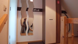 Une pièce avec une porte en bois, un mur blanc orné d’un miroir qui reflète une girafe et une personne, et une chaise en bois.