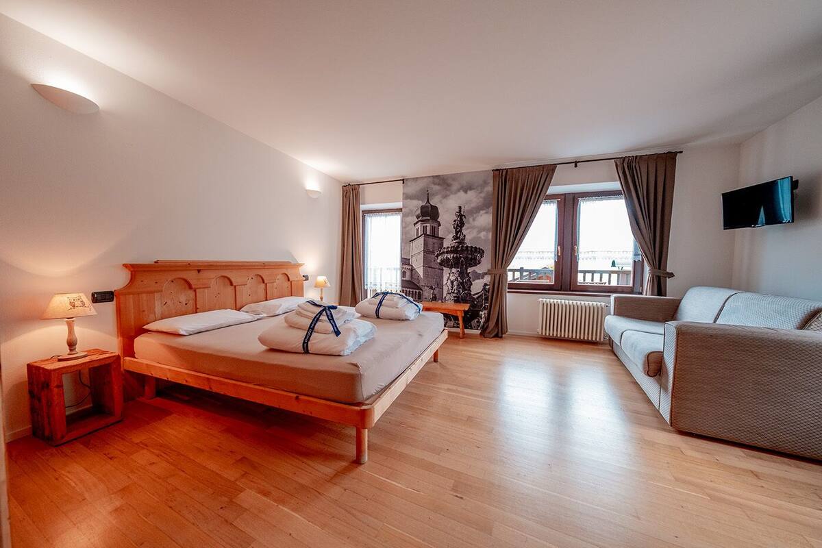 Habitación de hotel con una cama de madera, una mesita con una lámpara, un sofá y un amplio ventanal con cortinas.