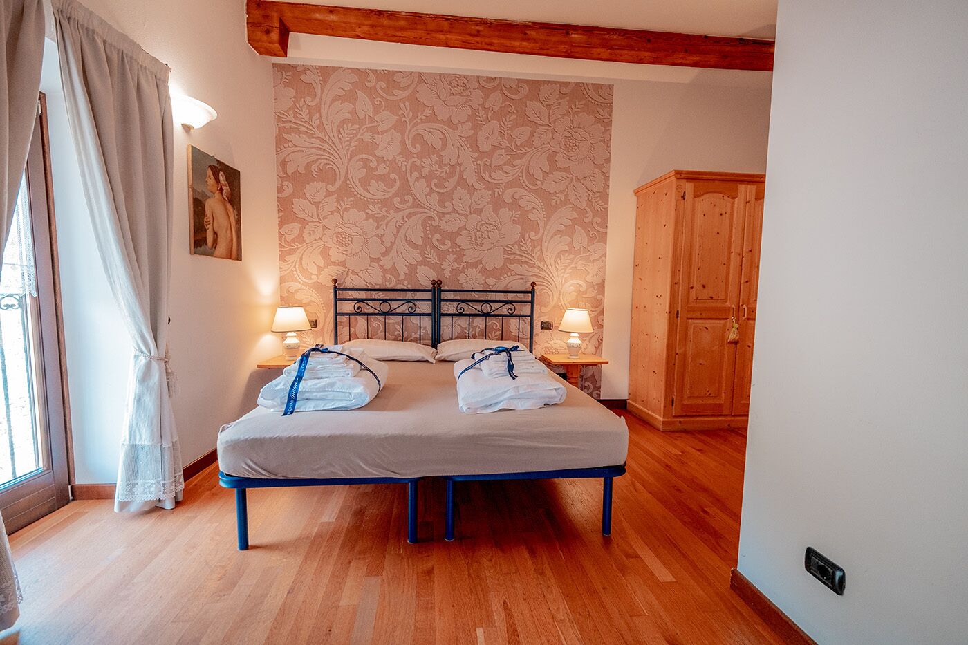 A neatly made bed with towels and a headboard, two bedside lamps, and a wooden wardrobe in a room with patterned wallpaper and wooden flooring.