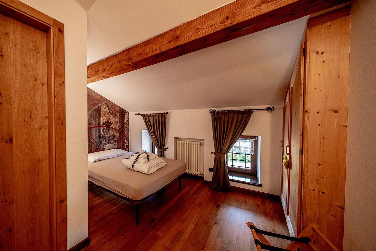 Un dormitorio con techo de madera, una cama con colchón blanco y una toalla doblada, una ventana con cortinas y un radiador.