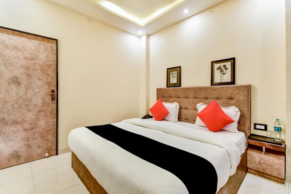 A hotel room with a large bed, a wooden headboard, a nightstand with a water bottle, and a door with a textured panel. - Hotel Frontier Royal (Kota)