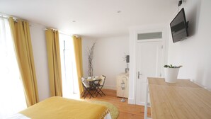 A bedroom with a bed, a wooden desk, a television, and a small table with a plant.
