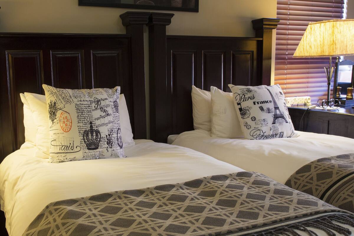 A hotel room with two beds, each with decorative pillows, a dark wooden headboard, and a lamp on a bedside table.