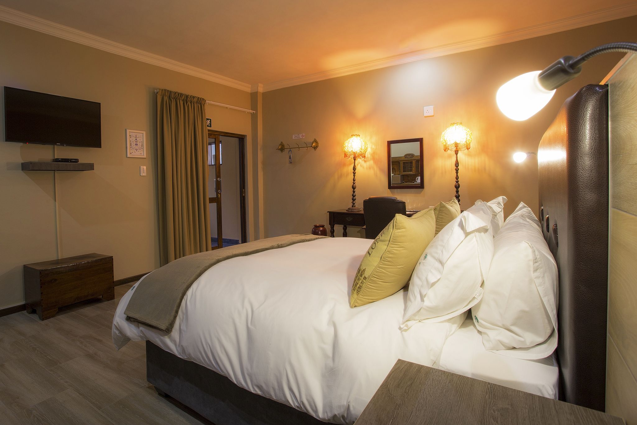 A hotel room with a large bed, bedside lamps, a flat-screen TV, a wooden bench, and a door with curtains.