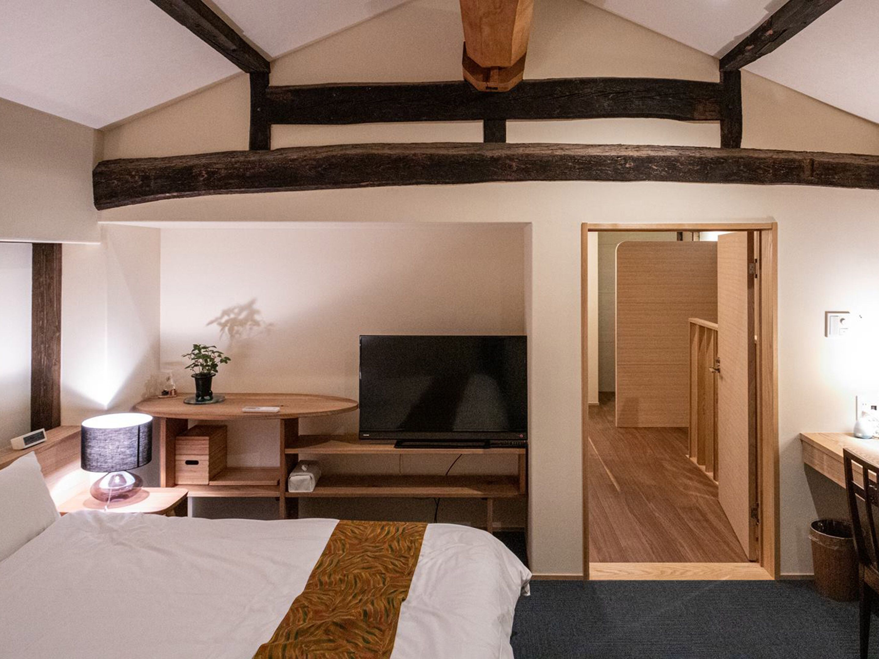 Una camera da letto con un letto, un comodino, una lampada, TV, scrivania, sedia e una porta che dà su un'altra stanza.