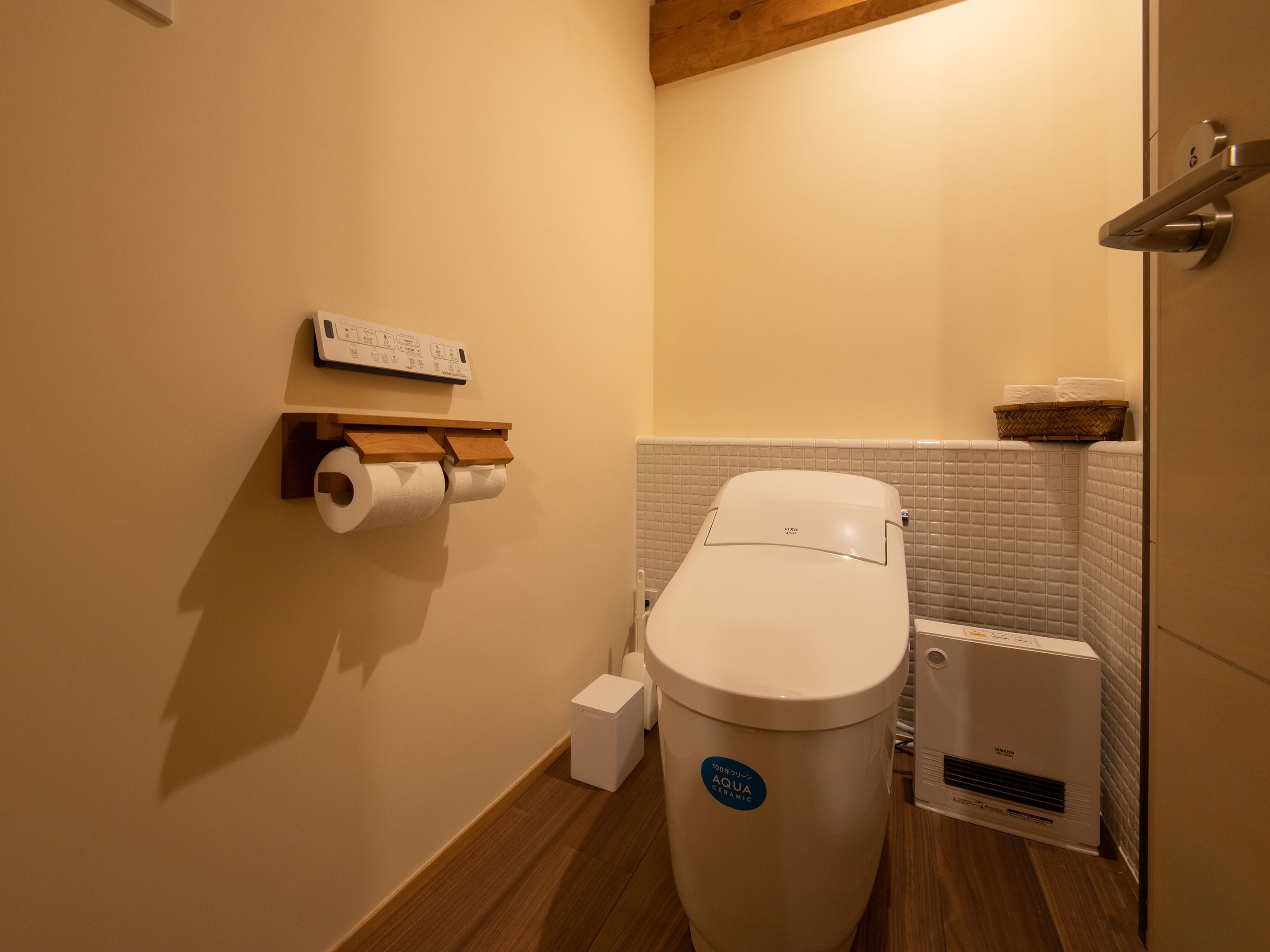 A compact bathroom with a toilet, a paper towel holder, a small trash bin, and a wall-mounted heater.