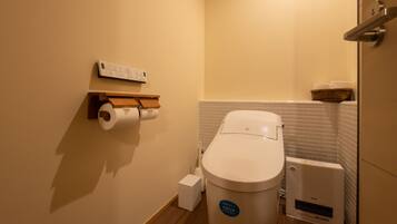 A compact bathroom with a toilet, a paper towel holder, a small trash bin, and a wall-mounted heater.