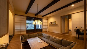 A traditional Japanese-style living room with tatami flooring, a low wooden table, and a large window with a view of a stone wall and plants. - UCHIKO-Inn HISA (Uchiko)