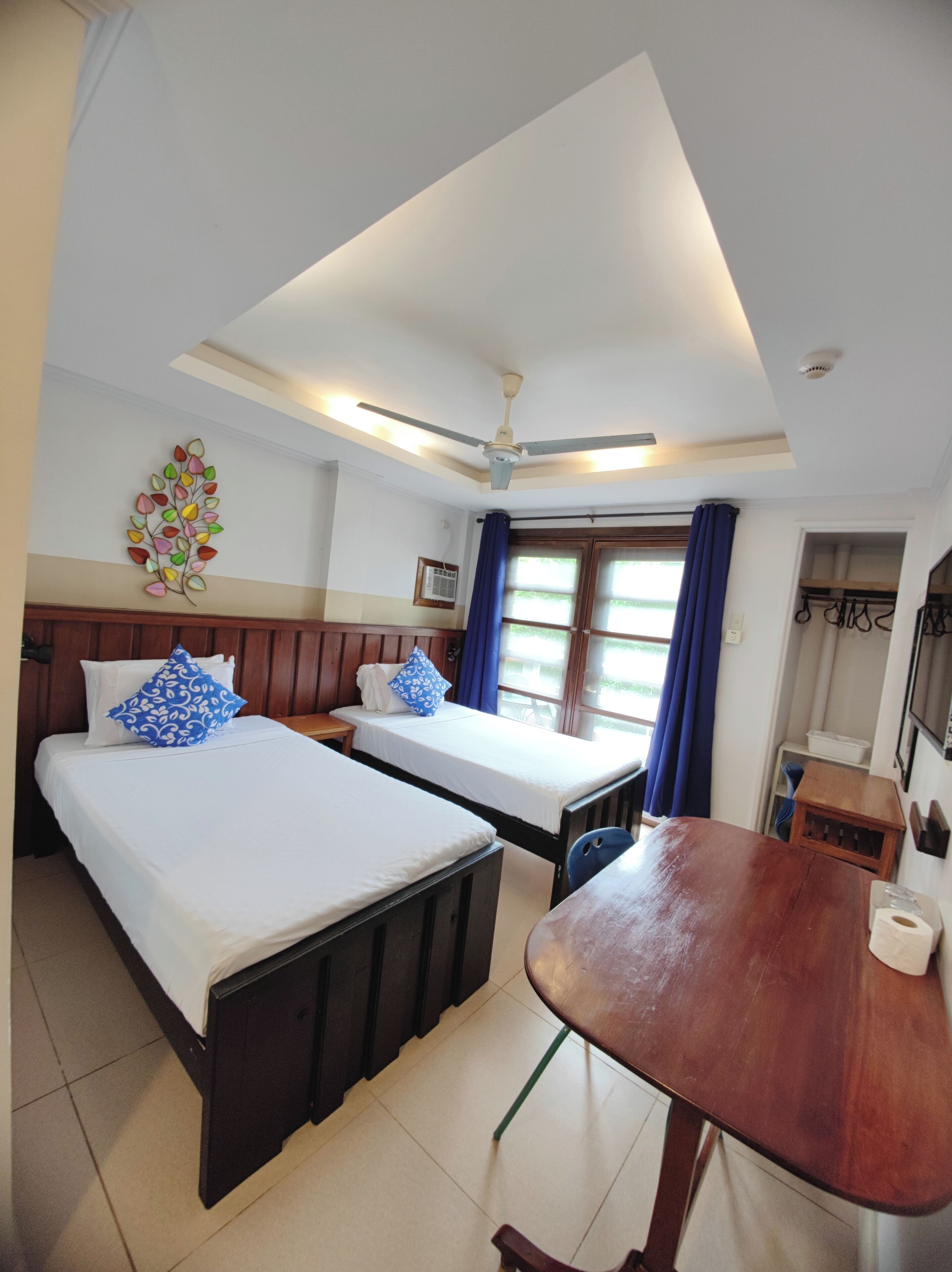 A hotel room with two beds, a ceiling fan, a window with curtains, a small table, and a wall-mounted air conditioning unit.