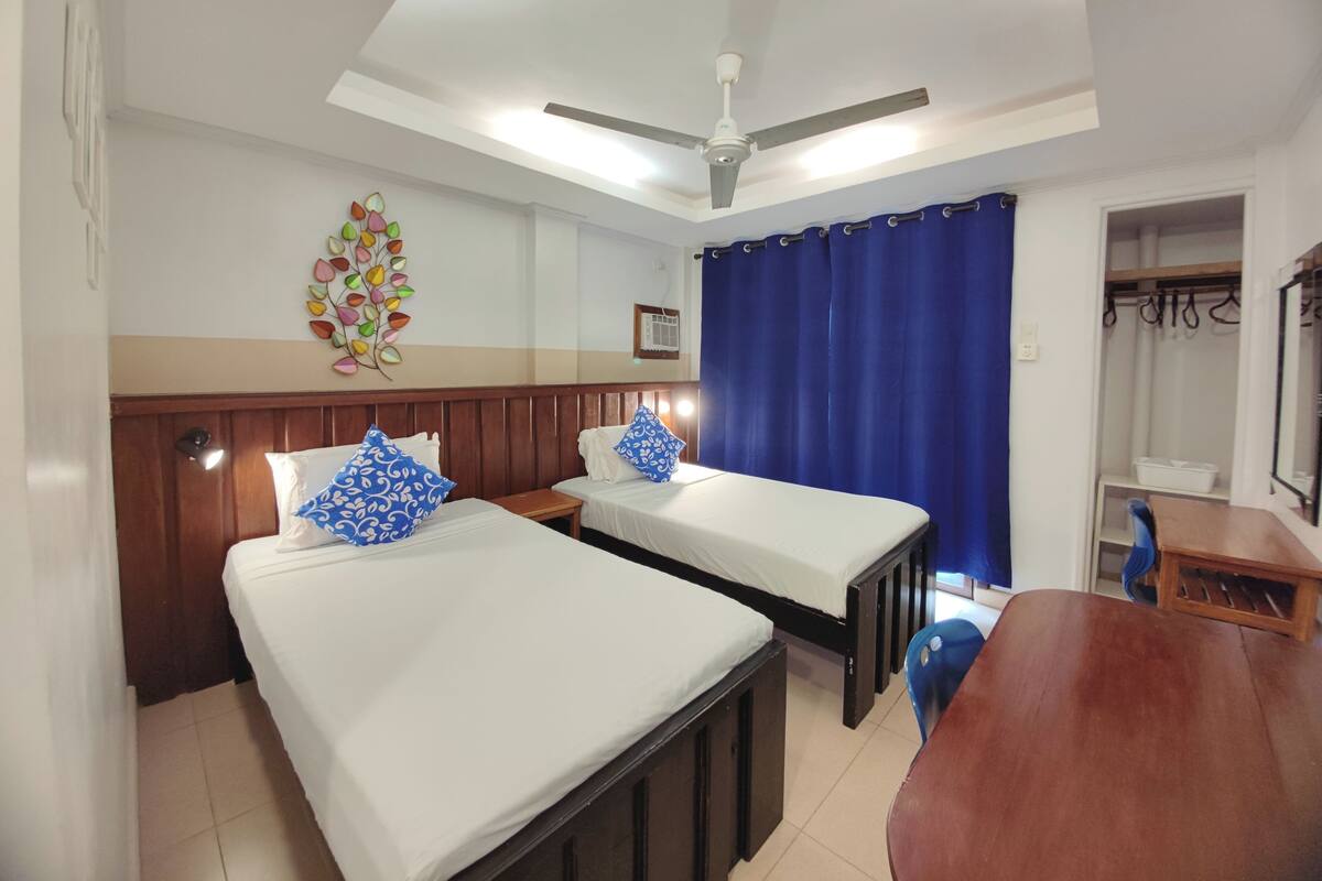 A hotel room with two beds, a wooden headboard, a ceiling fan, a blue curtain, and a small table.