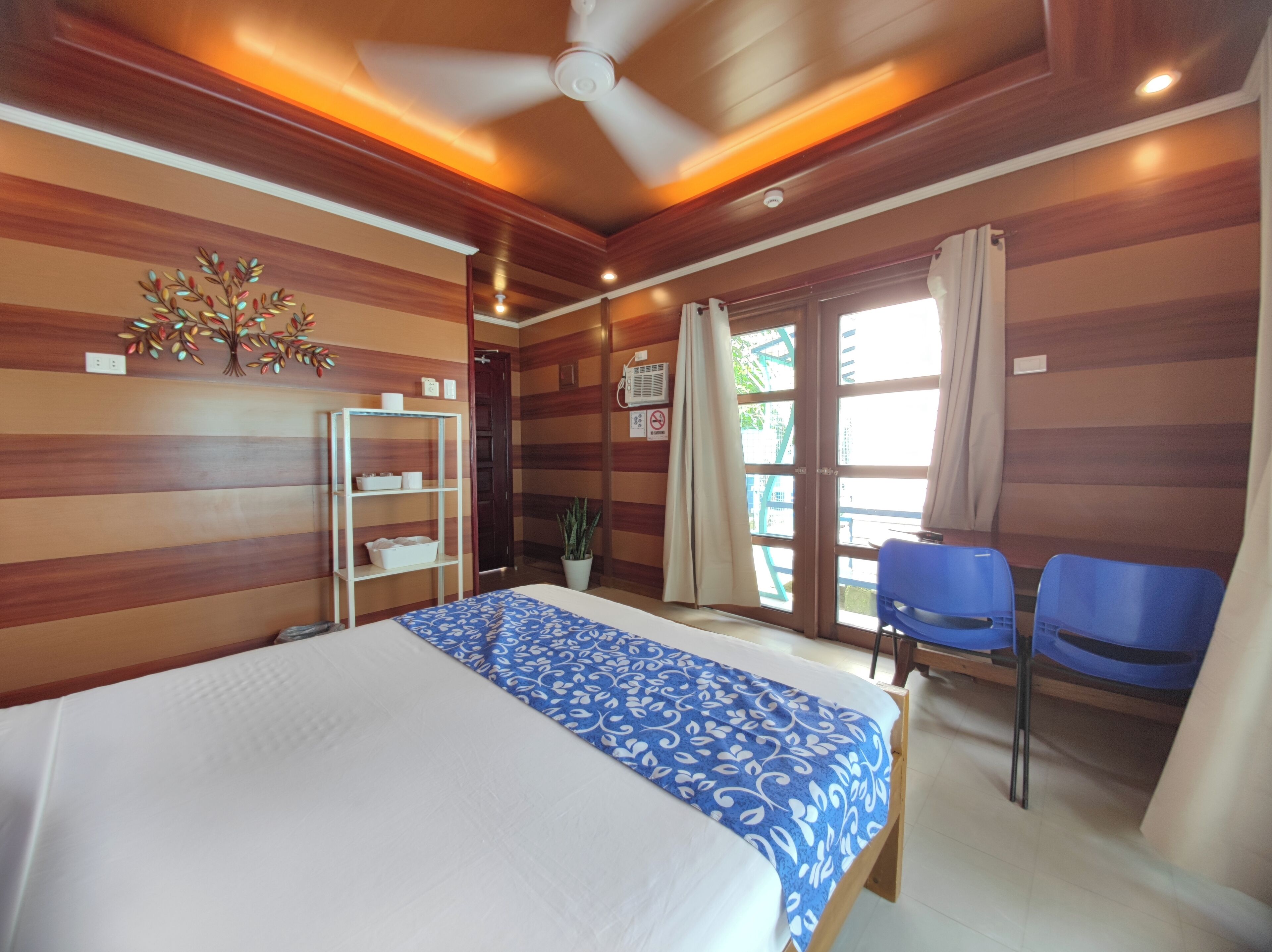 Una habitación de hotel con una cama, un ventilador de techo, una ventana con cortinas, una planta y una silla azul.