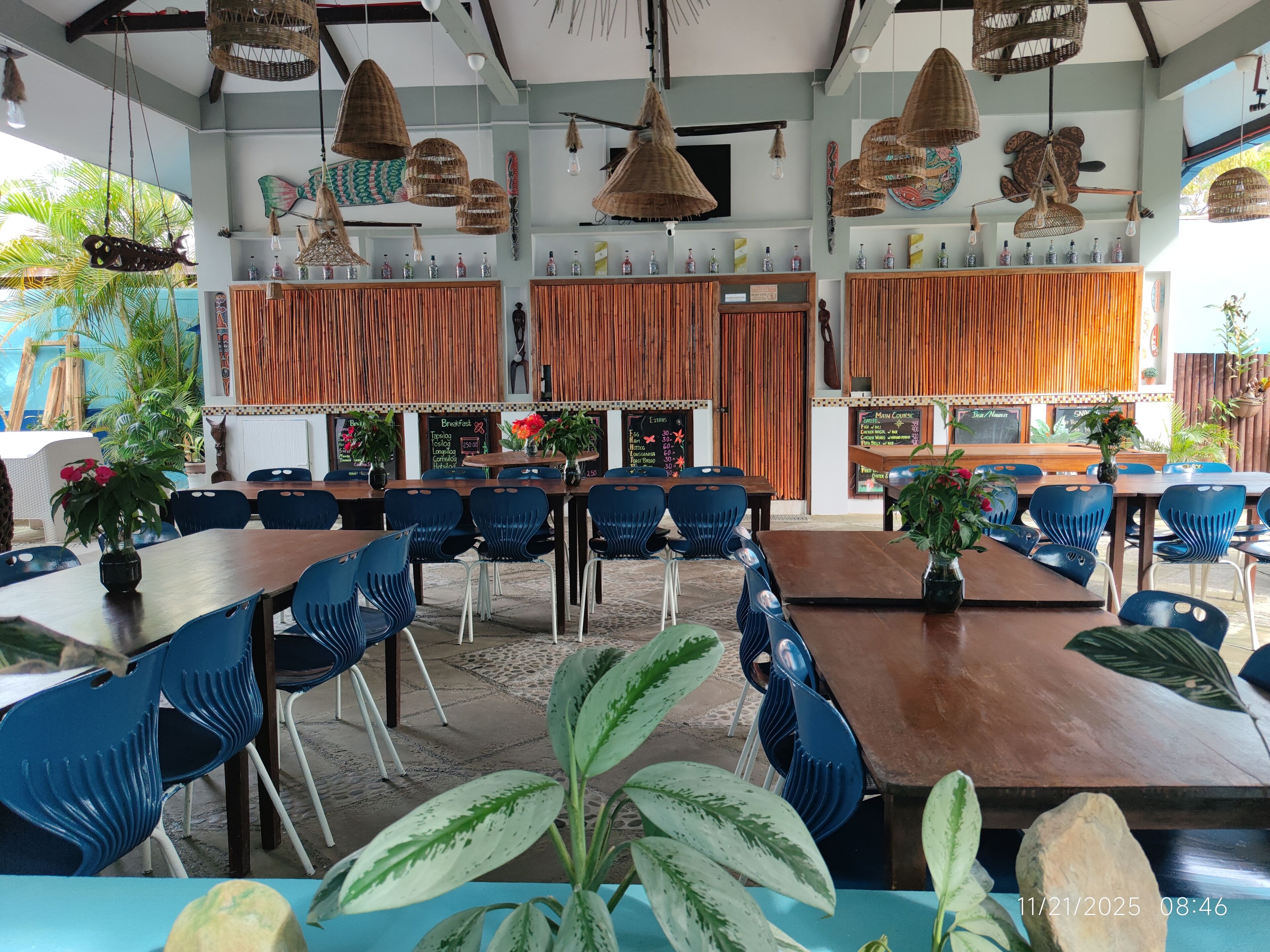 Ein Restaurant mit Holztischen und blauen Stühlen, daran hängen Körbchenlampen, und eine Speisekarte.