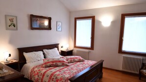 A bedroom with a wooden bed, white bedding, a nightstand with a lamp, and two windows with blinds. - Agriturismo Memento (Follina)