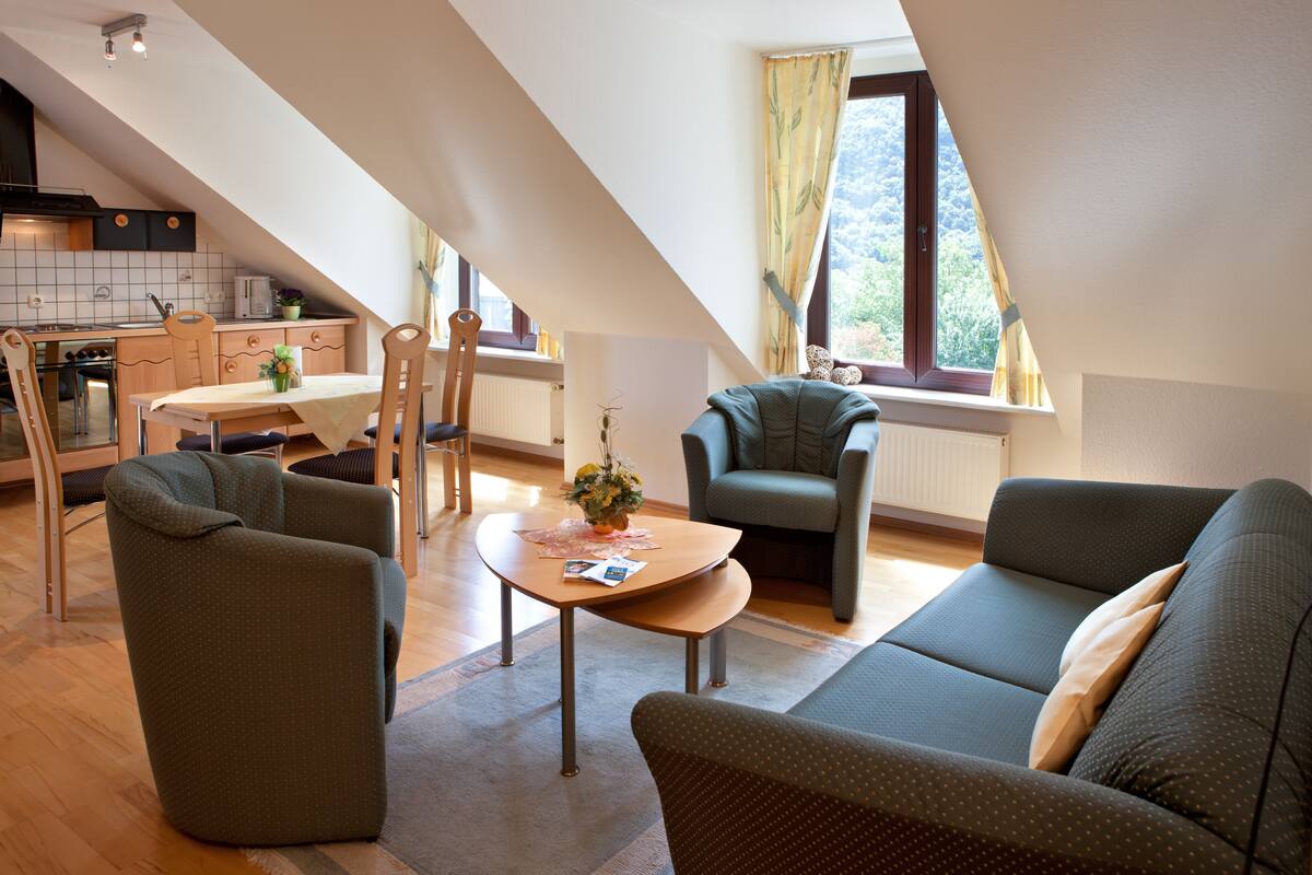 A modern living room with a sofa, armchairs, and a coffee table. There is a kitchen area with a stove and cabinets. A window with curtains is visible, and the room has a sloped ceiling.