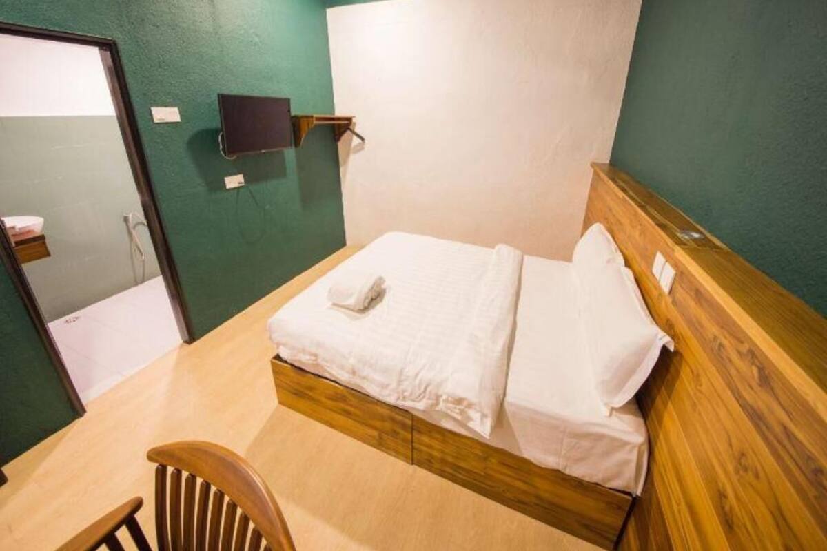 A single bed with white linens, a wooden headboard, a small wooden chair, and a flat-screen TV mounted on the wall.